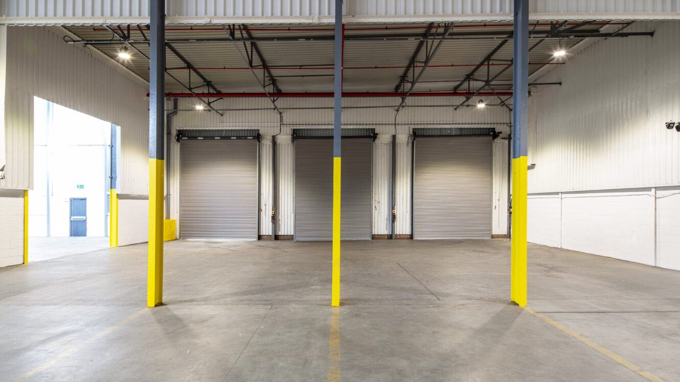 Interior of an empty warehouse with three closed roller shutters, yellow bollards, high ceiling, and visible overhead lighting.