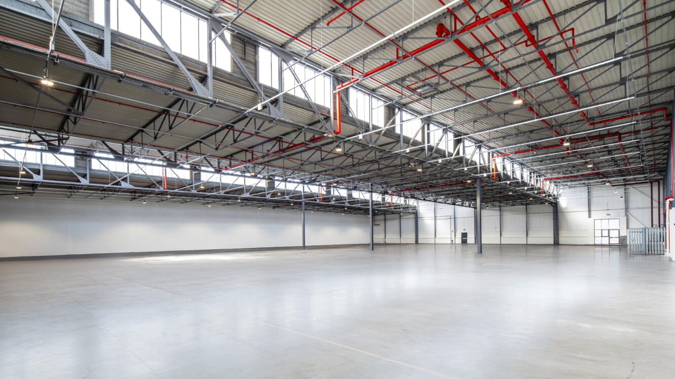Large empty industrial warehouse with high ceilings, exposed beams, skylights, and visible red pipework, illuminated by natural and artificial light.