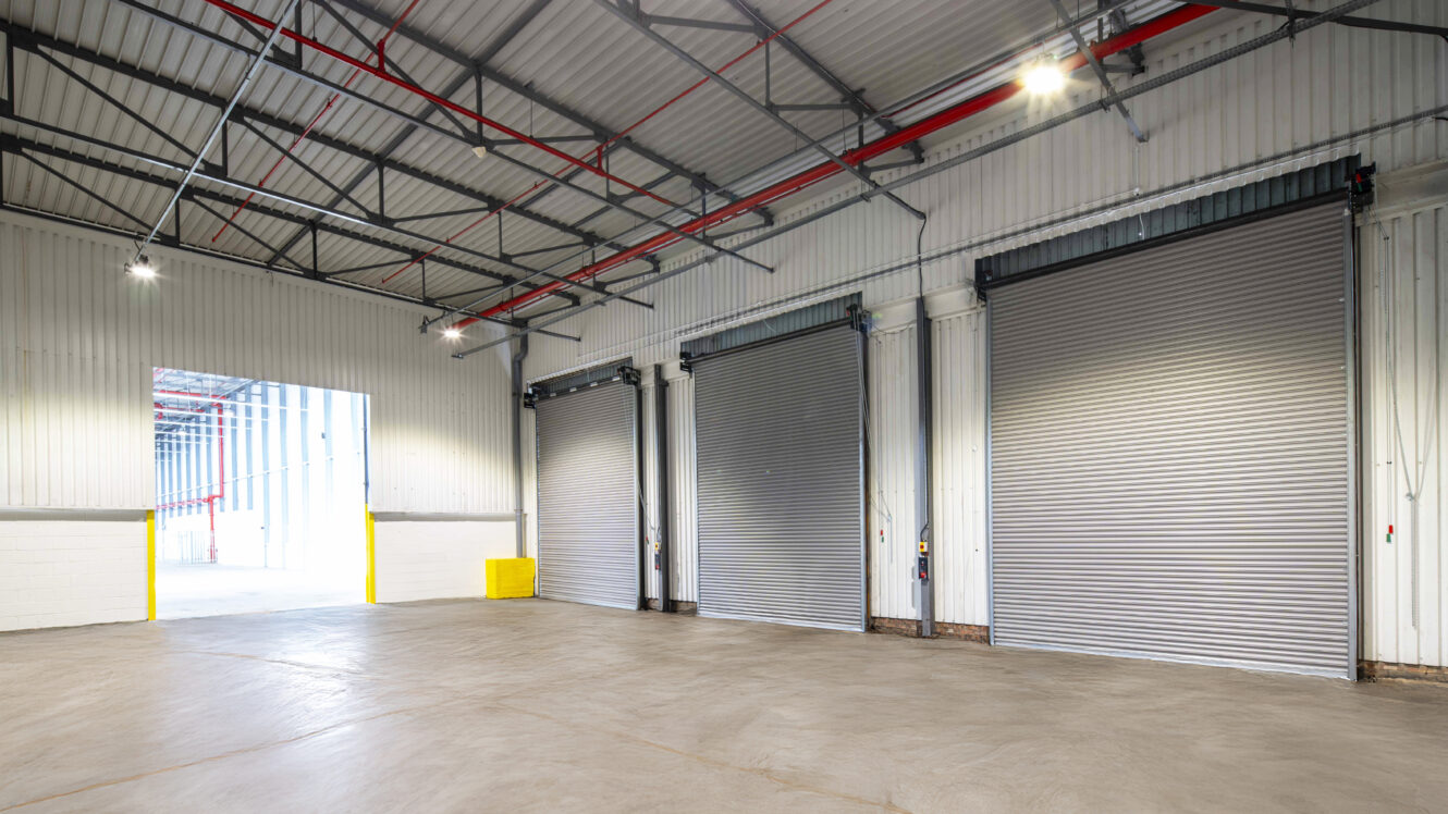 Interior of an empty industrial warehouse with three large closed roller shutter doors and an open entryway to the left. The space has concrete floors and a high metal ceiling.