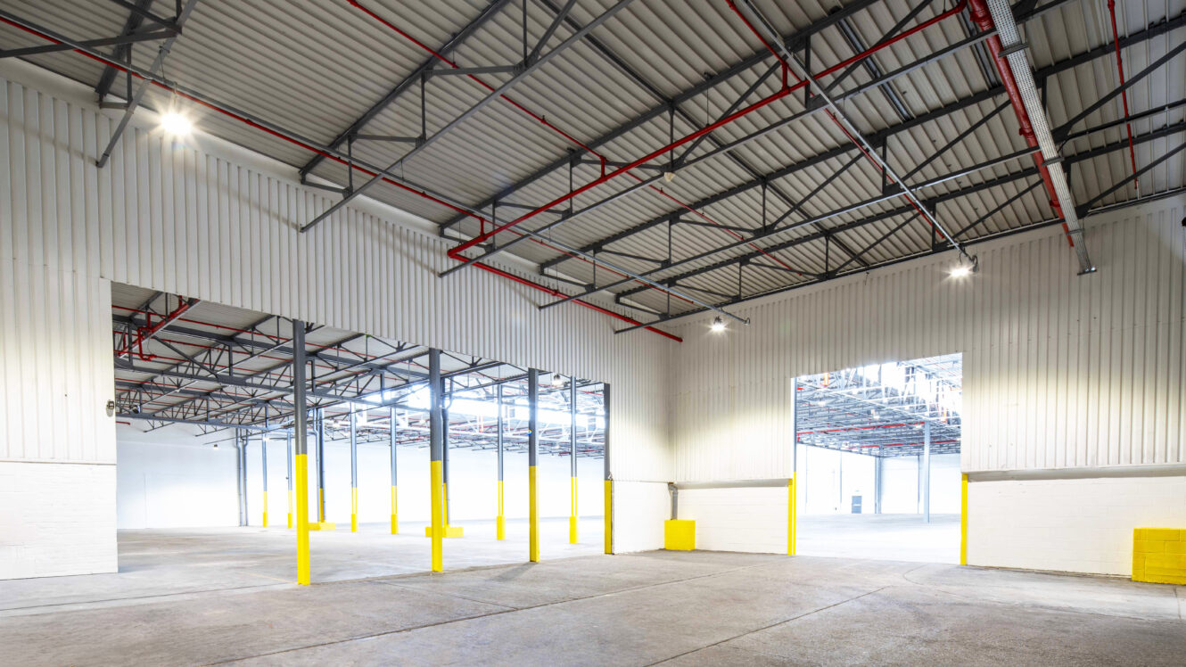 Interior of a large, empty warehouse with high ceilings, exposed beams, metal roof, yellow column guards, and bright overhead lighting.