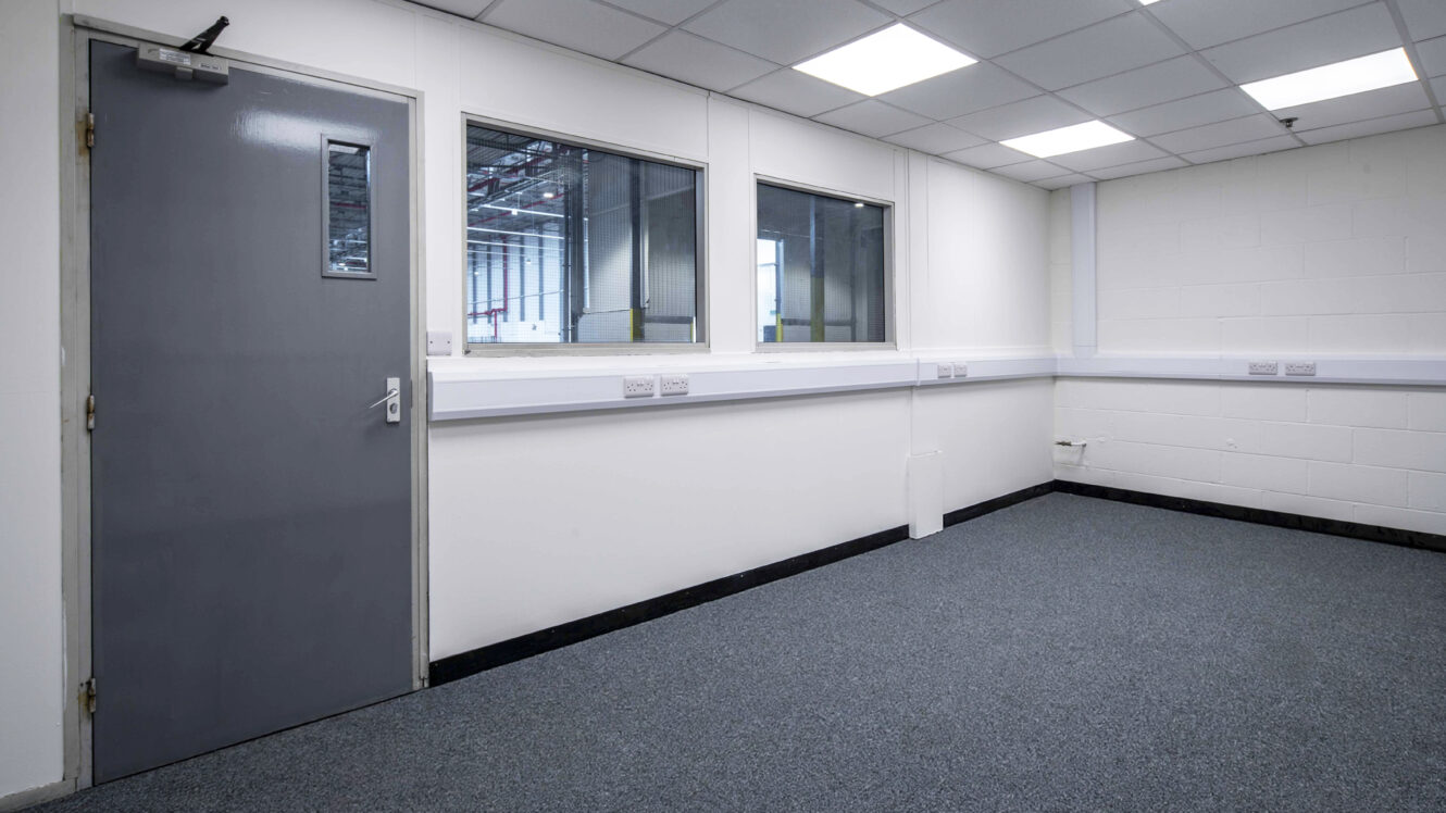 Empty office room with grey carpet, white walls, two internal windows, a grey door with a small window, and a grid ceiling with fluorescent lights.