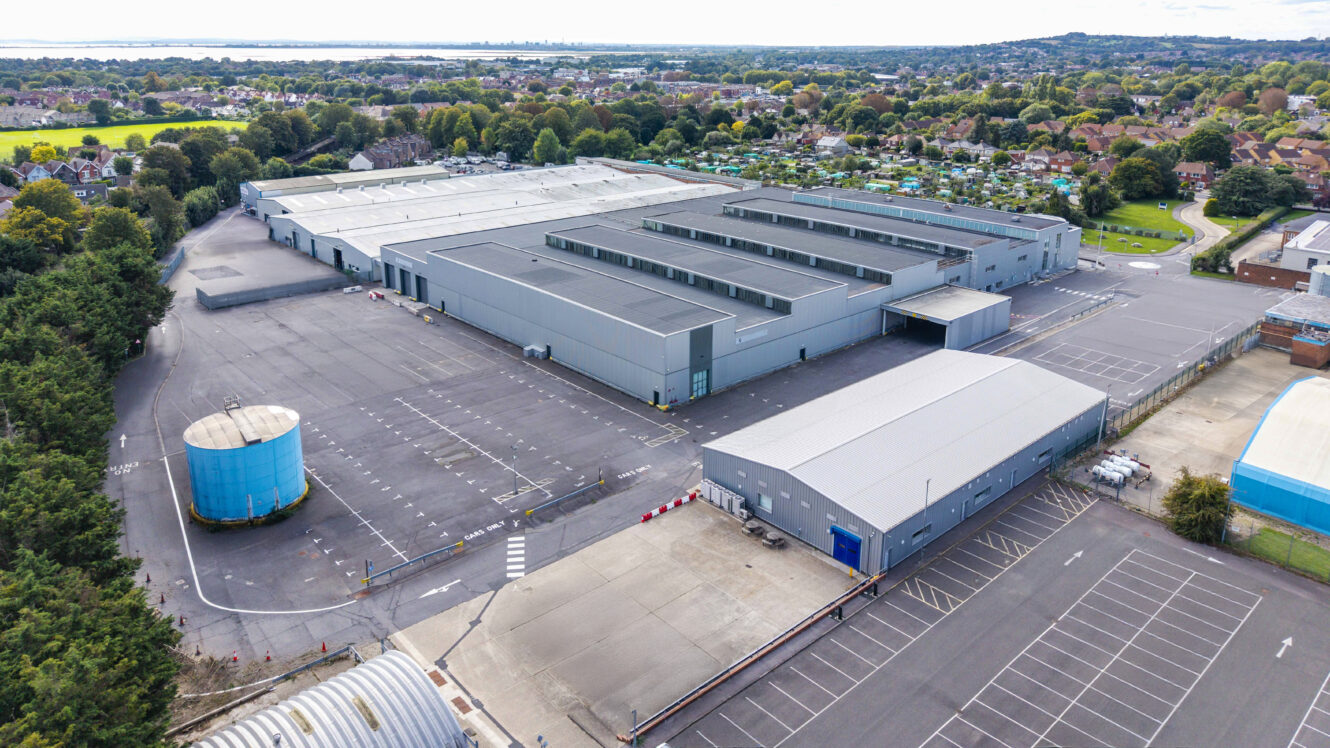 Aerial view of a large industrial warehouse complex with adjoining car parks and a blue cylindrical tank, surrounded by residential and green areas.