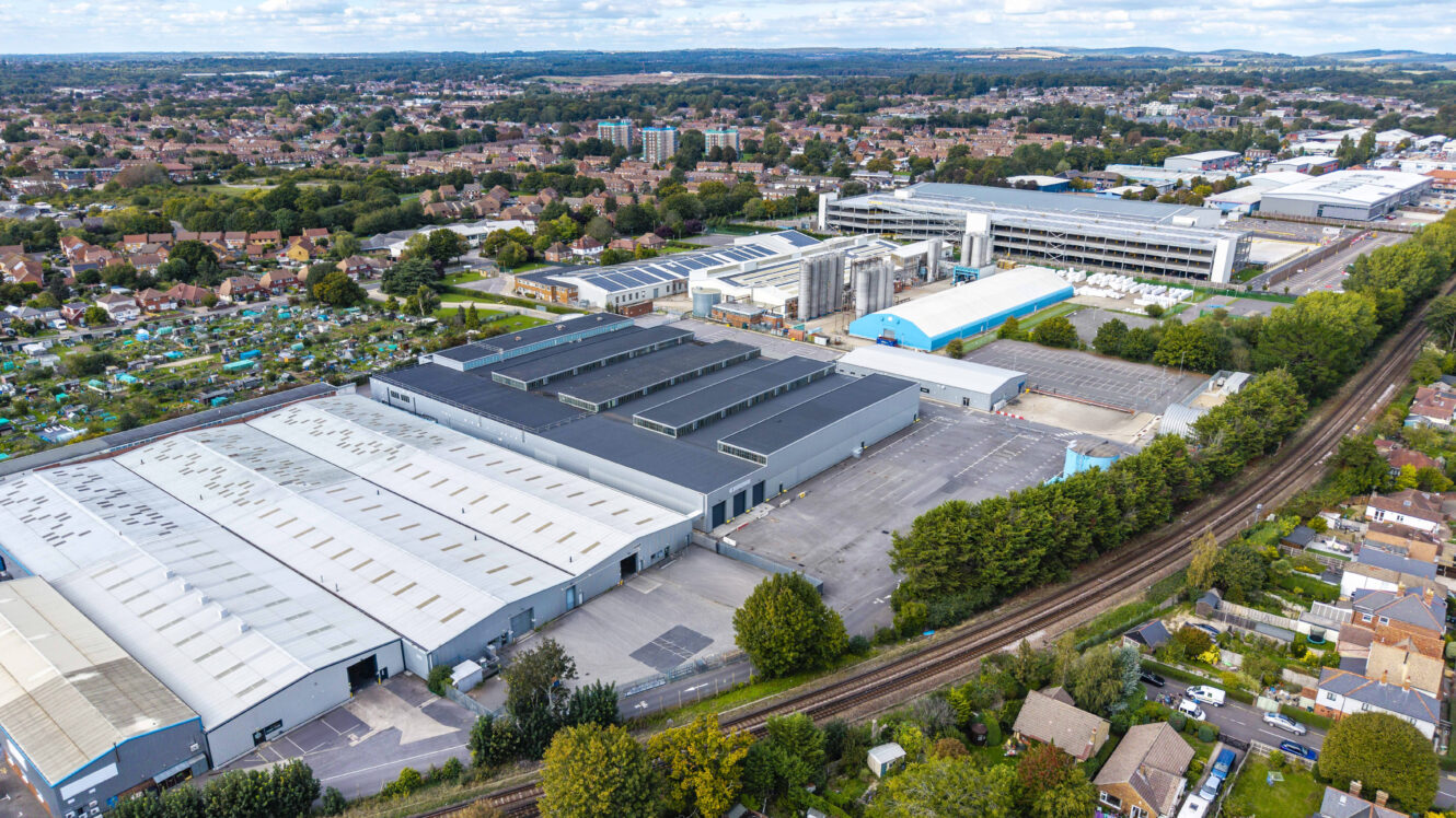 Aerial view of a large industrial complex with multiple warehouse buildings, surrounded by residential houses, trees, and a railway line.