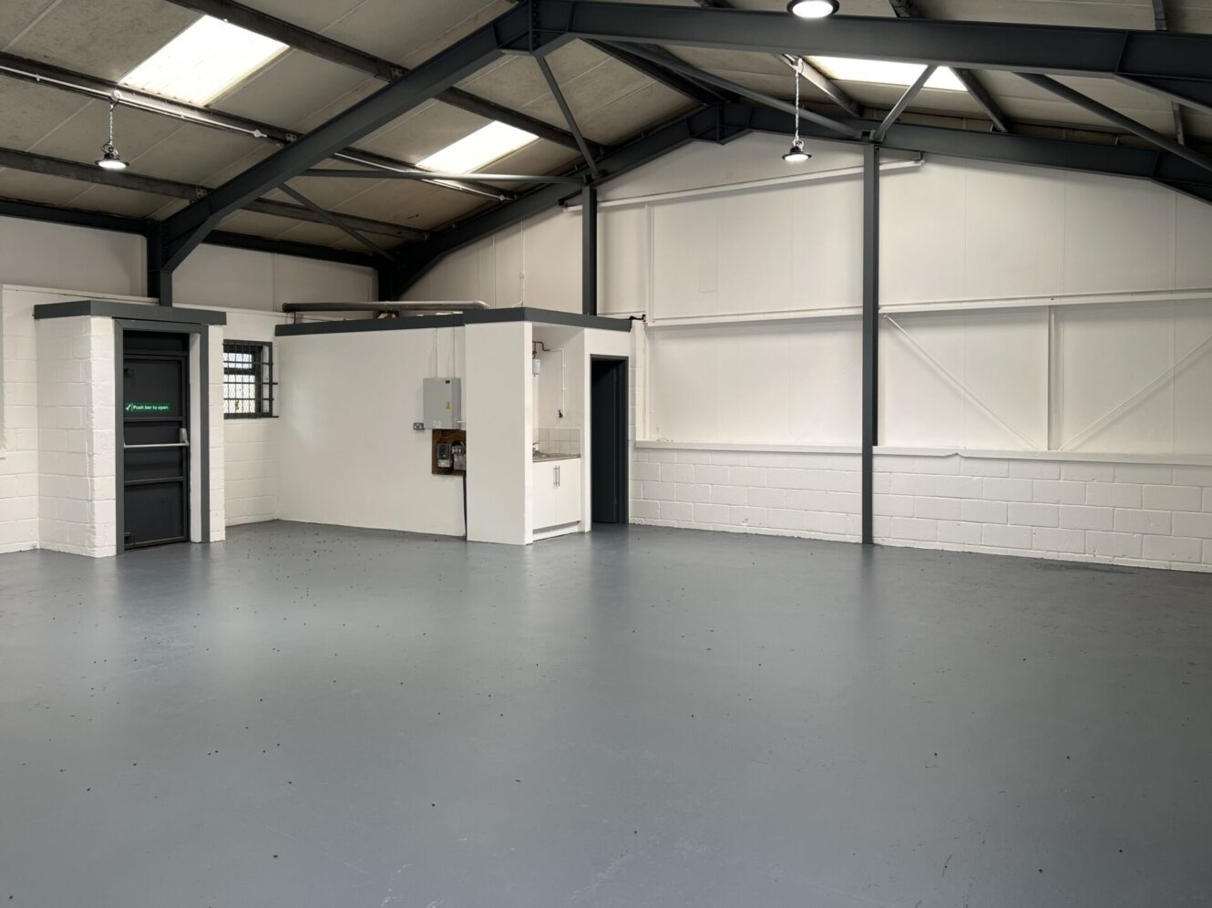 Interior of an empty industrial warehouse with grey flooring, white brick walls, high ceiling, exposed beams, and a small enclosed office area.