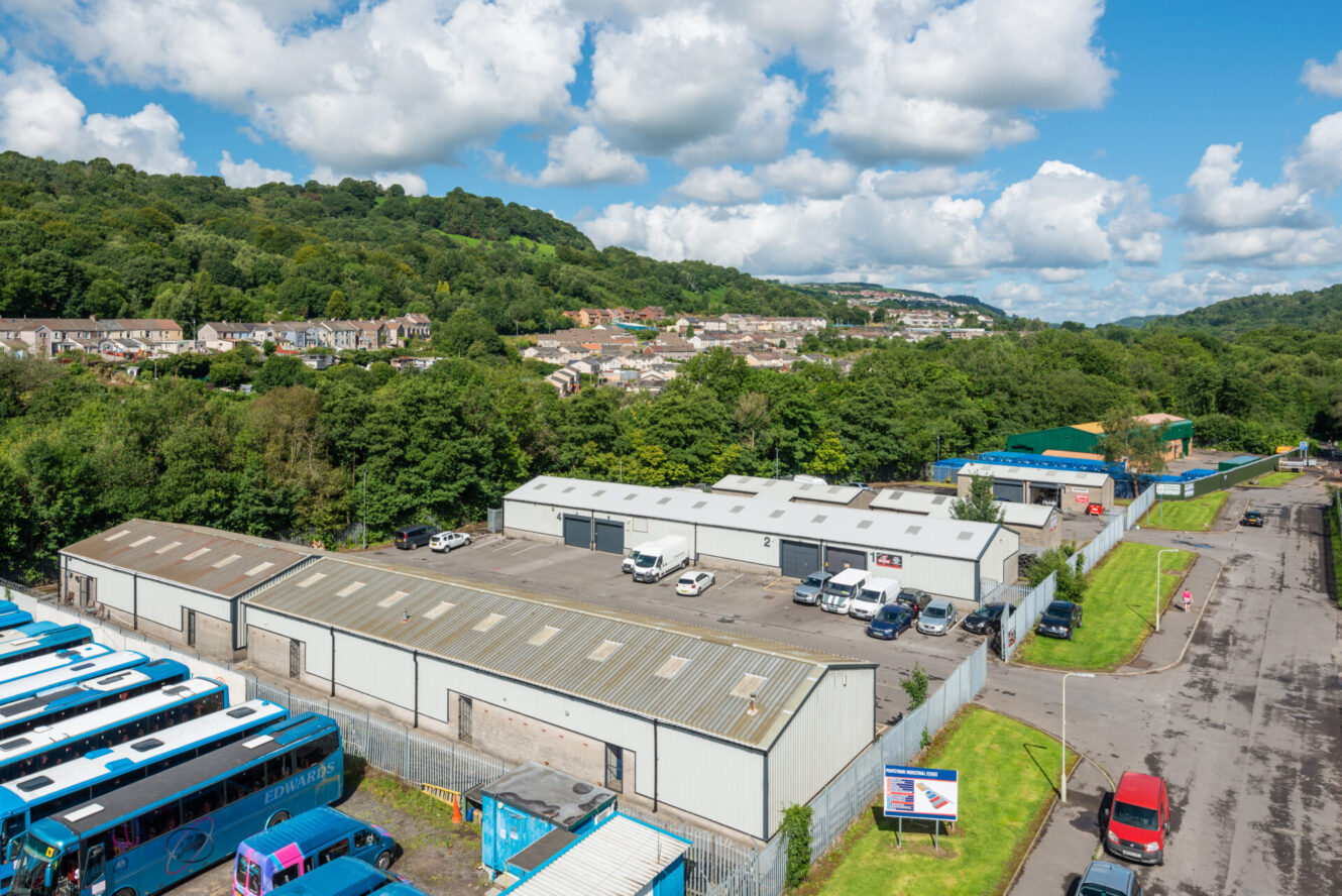 Aerial view of a commercial industrial estate with warehouses, parked vehicles, nearby buses, trees, and a hillside residential area in the background under a partly cloudy sky.