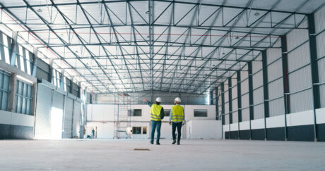 Two people wearing safety vests and helmets walk inside a large, empty warehouse with high ceilings and steel girders.