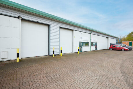 A row of industrial warehouse units with white roller shutters, yellow and black bollards, and a red car parked outside on a paved car park.