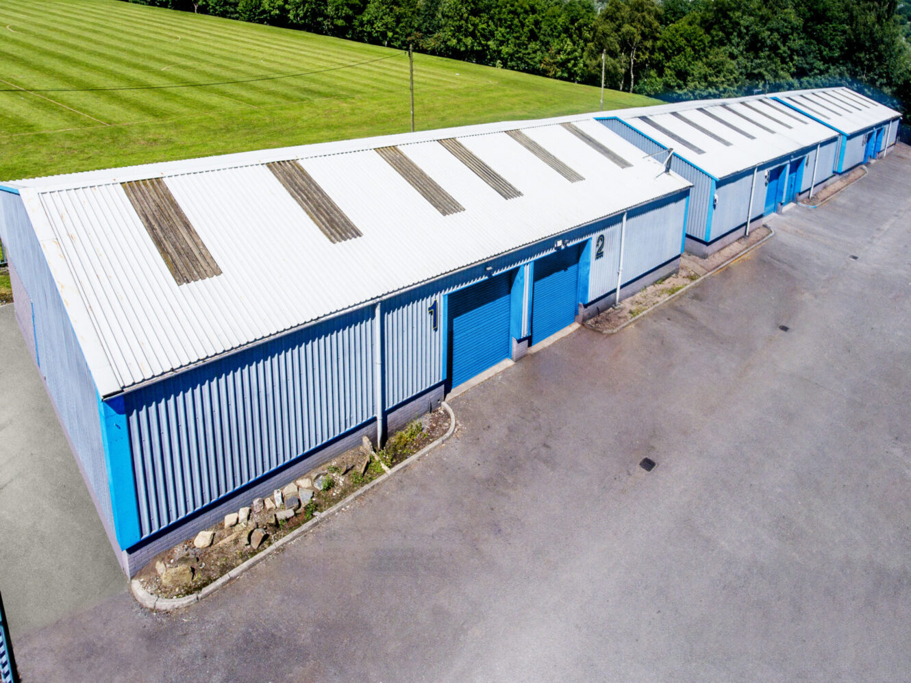 A row of blue and white industrial storage units with closed garage doors, next to an empty paved area and a grass field in the background.