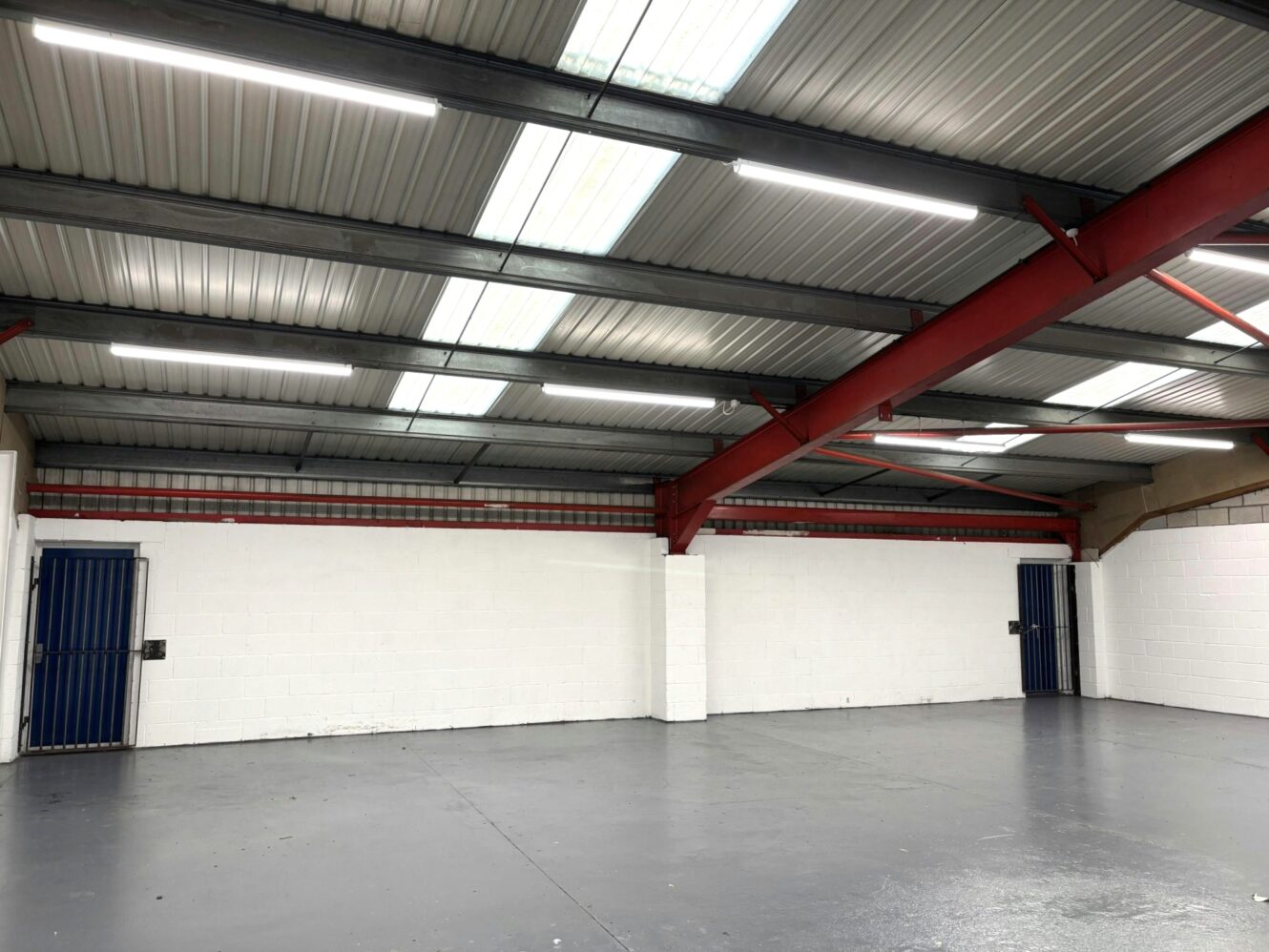 Empty industrial warehouse with a grey concrete floor, white brick walls, blue metal doors, and exposed red steel beams under a corrugated metal roof with strip lighting.
