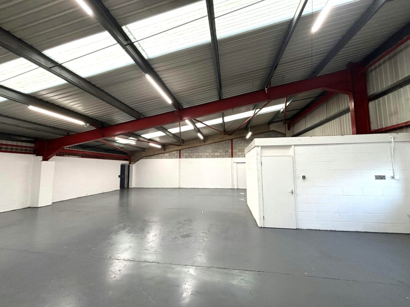 Empty industrial warehouse space with high ceilings, red steel beams, concrete floors, white brick walls, and a small enclosed office area in the corner.