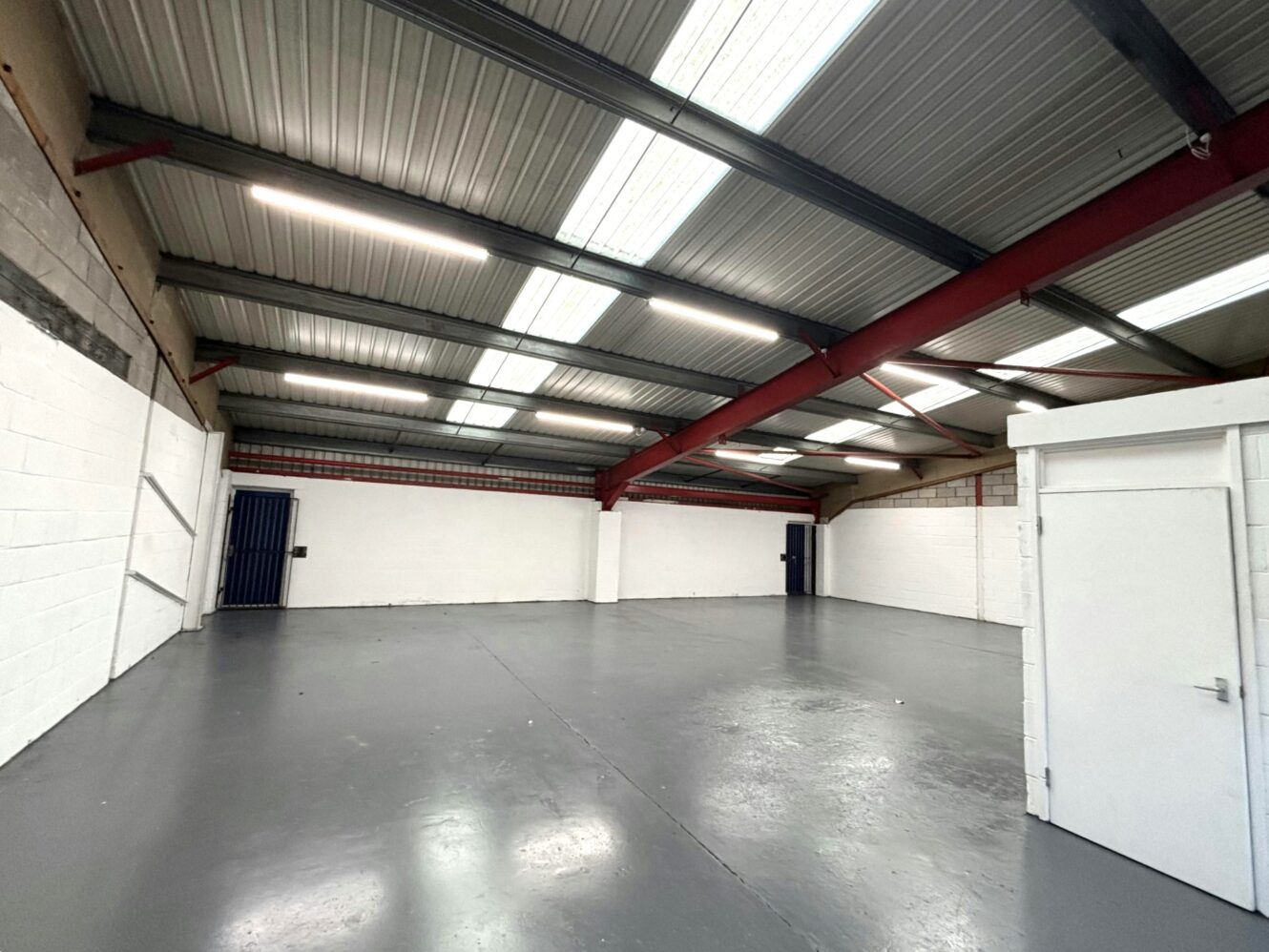 Interior of an empty industrial warehouse with grey floors, white walls, metal ceiling, red support beams, and a small enclosed office in the corner.