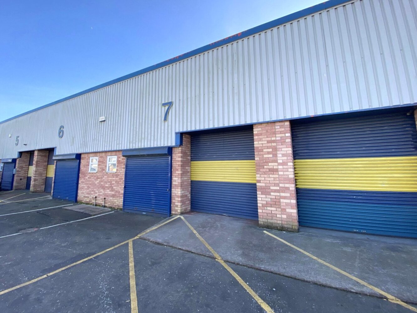Row of industrial storage unit doors numbered 5, 6, and 7, with yellow and blue shutters, brick walls, and marked parking bays in front.