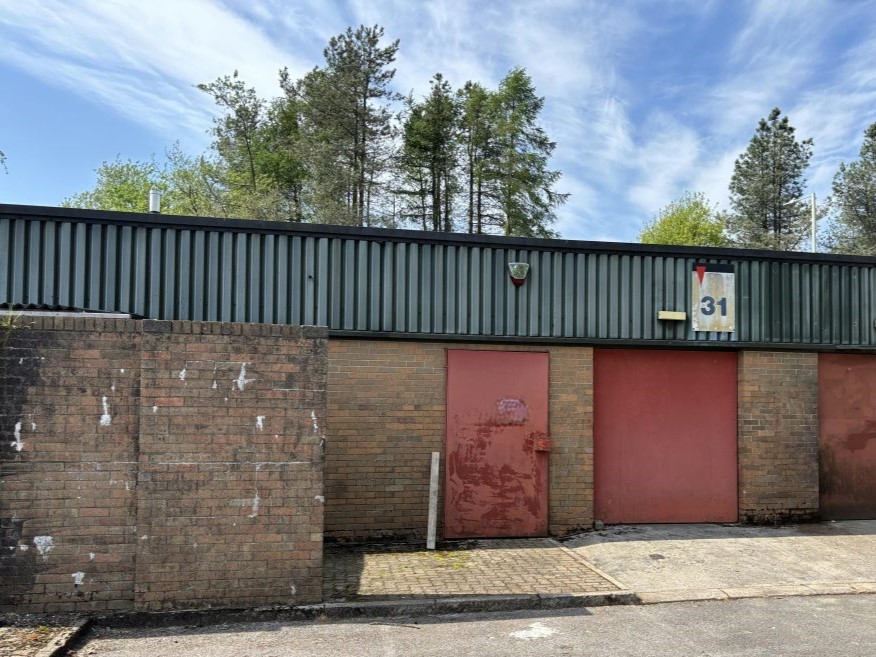 A brick industrial building with two large red doors and a sign marked 31 under a partly cloudy sky, surrounded by tall trees in the background.