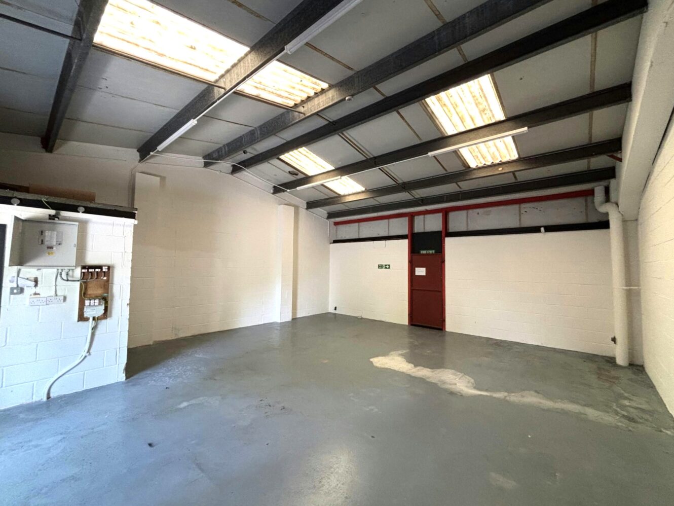 Empty industrial warehouse unit with concrete floor, white painted brick walls, exposed beams, fluorescent lights, and a red door with windows along the upper part of the wall.