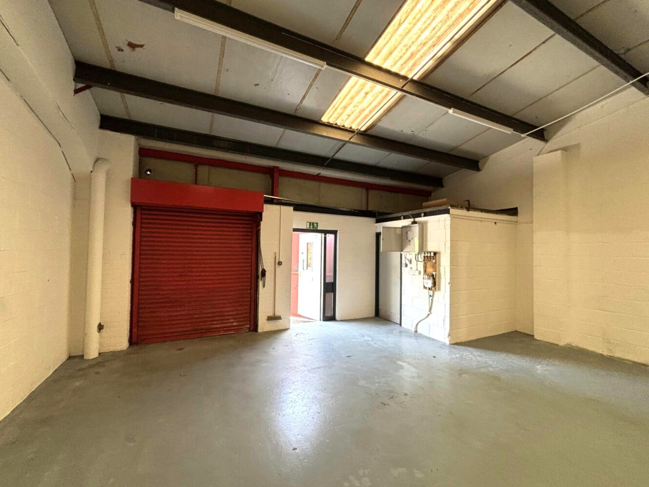 Empty industrial unit with white walls, grey concrete floor, red metal roller shutter door, overhead fluorescent lights, and exposed wiring on a small partitioned area.