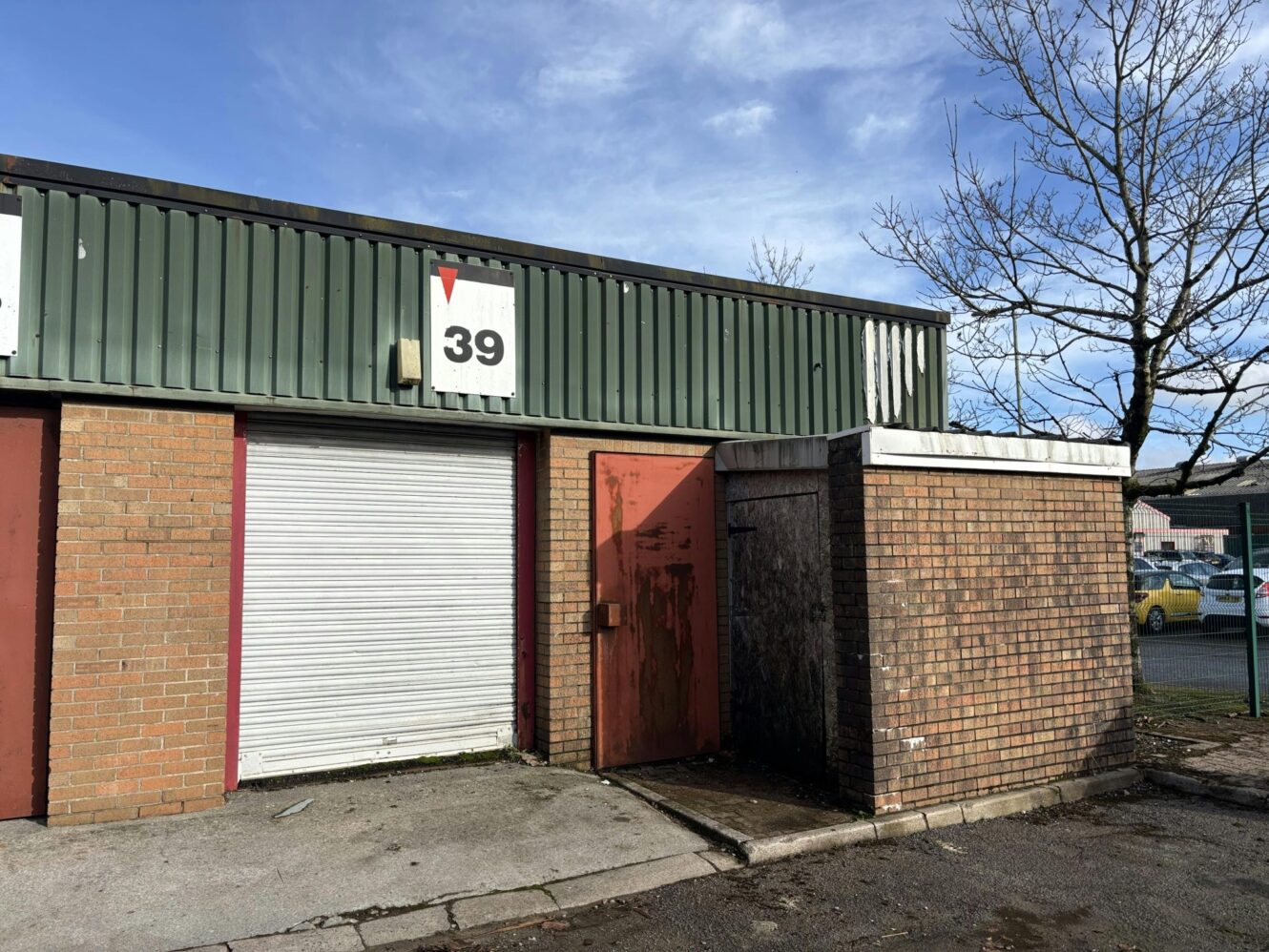 Single-storey industrial unit with a corrugated metal facade, a closed white roller shutter door, red metal door, and a sign displaying the number 39 above the entrance.