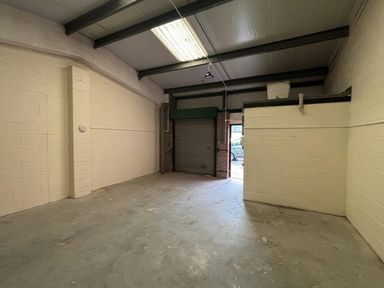 Empty industrial unit with high ceiling, concrete floor, white brick walls, exposed beams, and partially open metal roller shutter letting in natural light.