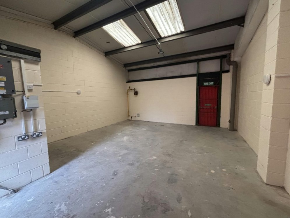 Empty industrial room with white brick walls, exposed ceiling beams, fluorescent lights, and a red exit door at the back. Concrete floor and electrical panels are visible.