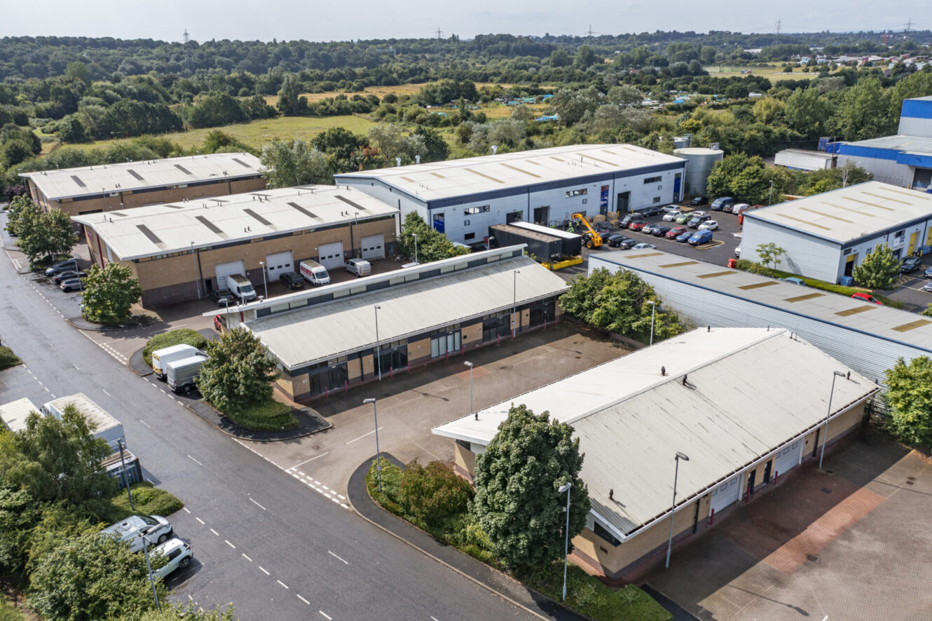 Aerial view of an industrial estate with several warehouses, loading bays, parked vehicles, and surrounding greenery on a clear day.