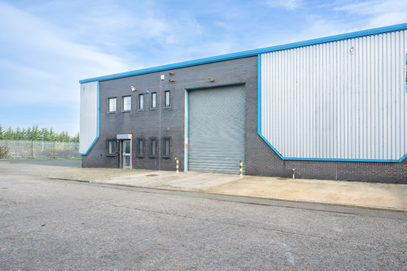 A large industrial warehouse with a brick façade, metal cladding, small windows, an entrance door, and a closed roller shutter garage door on a concrete yard.
