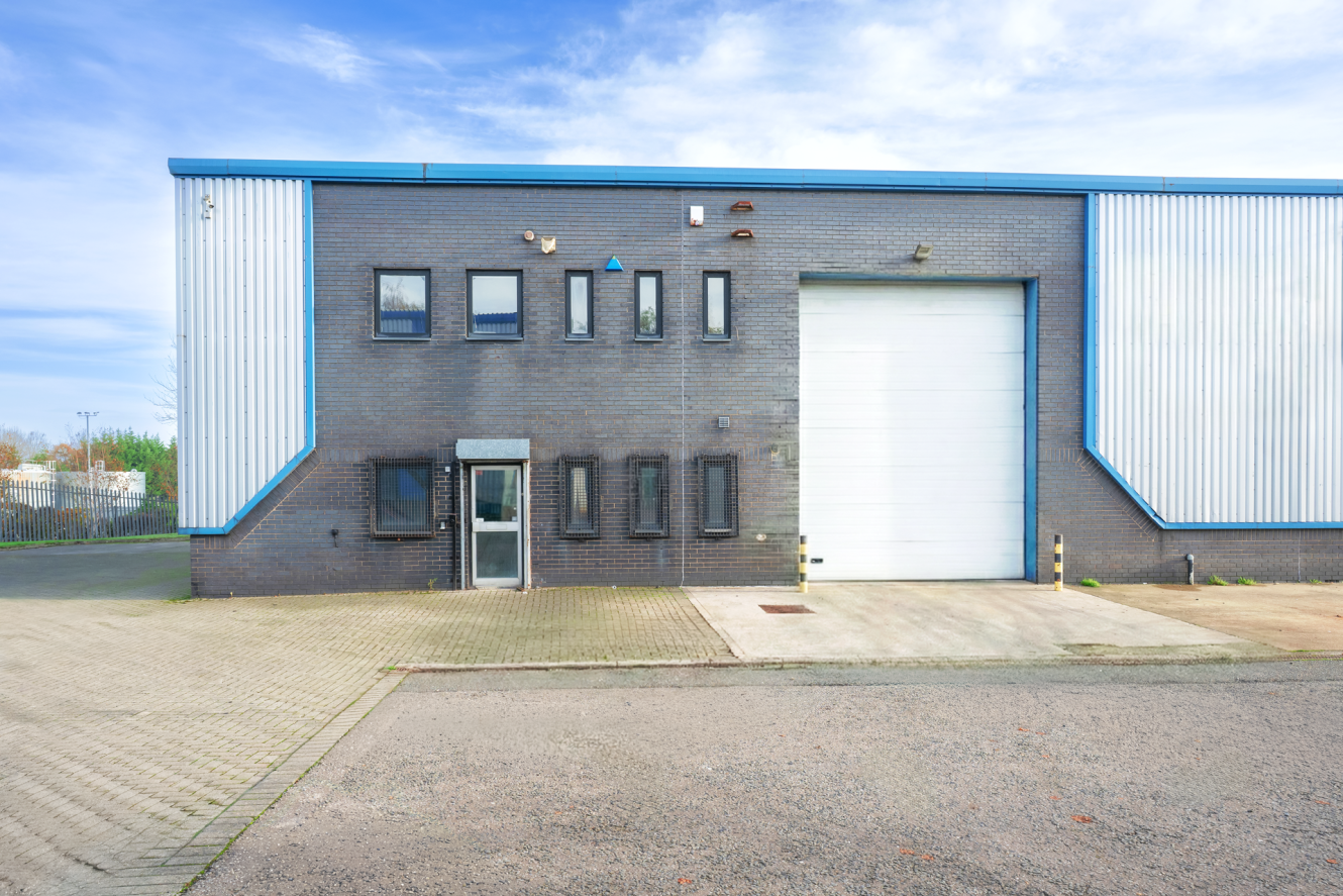 A modern industrial warehouse with a large white garage door, multiple small windows, and a single entrance door, set on a paved car park under a partly cloudy sky.