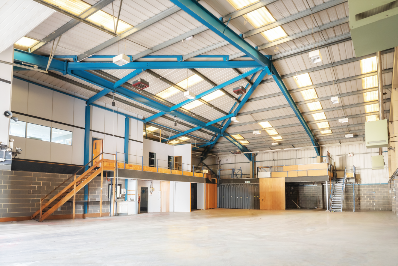 Spacious industrial warehouse interior with high ceilings, exposed beams, office areas, and metal staircases, illuminated by overhead lighting.