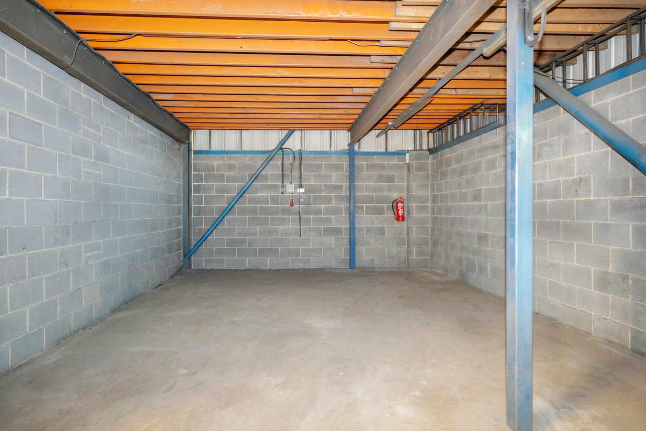 Empty industrial storage room with bare concrete floor, breeze block walls, exposed ceiling beams, and a red fire extinguisher mounted on the wall.