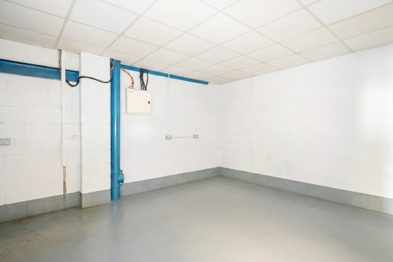 An empty, clean room with white-painted brick walls, grey floor, a tiled ceiling, and exposed blue pipes along the walls.