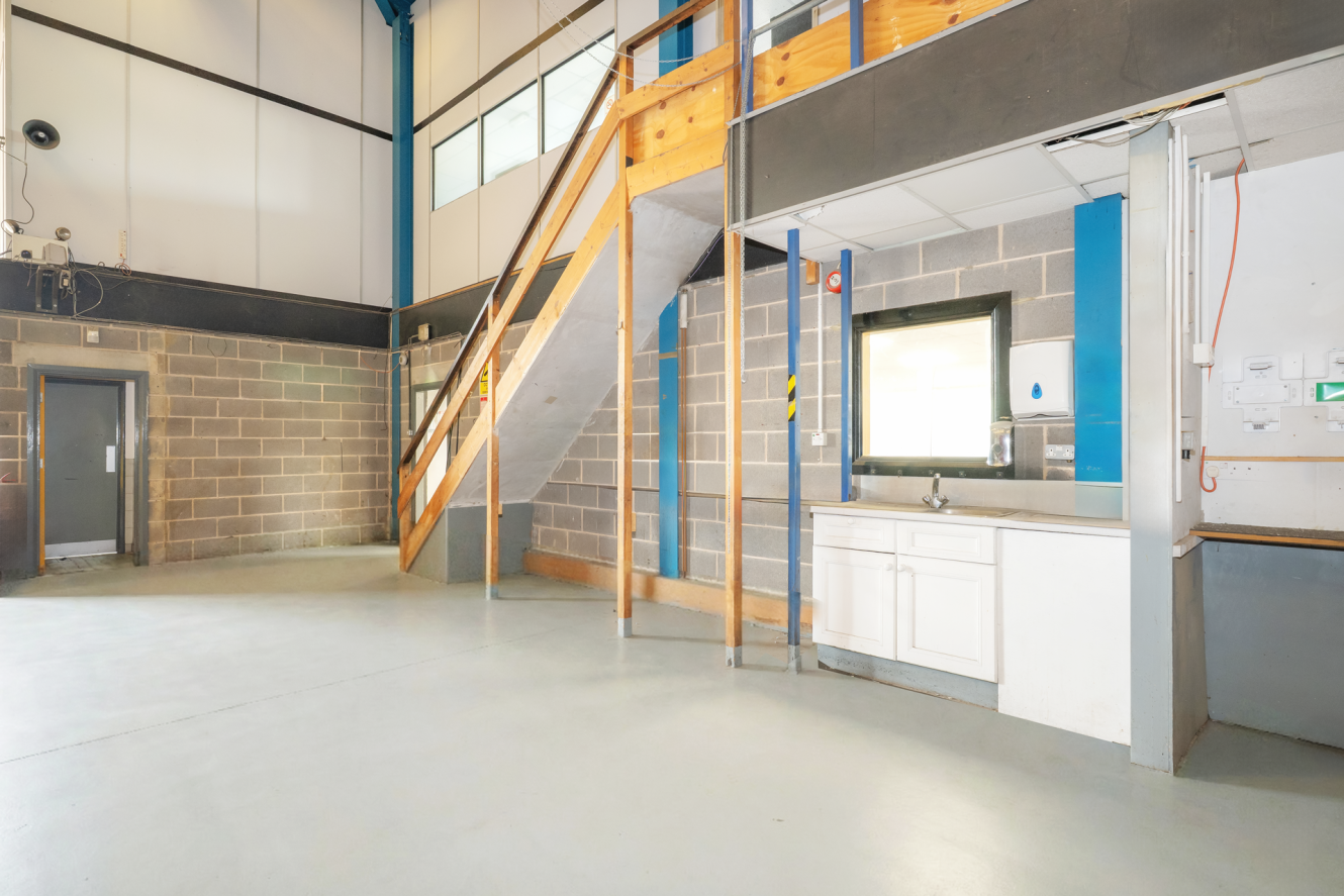 Interior of an industrial space with concrete floors, brick walls, a wooden staircase to a mezzanine, a window, a wall-mounted basin, and visible utility fittings.