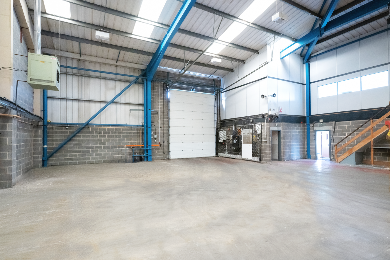 An empty industrial warehouse with concrete floors, brick walls, a large white roller shutter, metal beams, stairs, and windows letting in natural light.