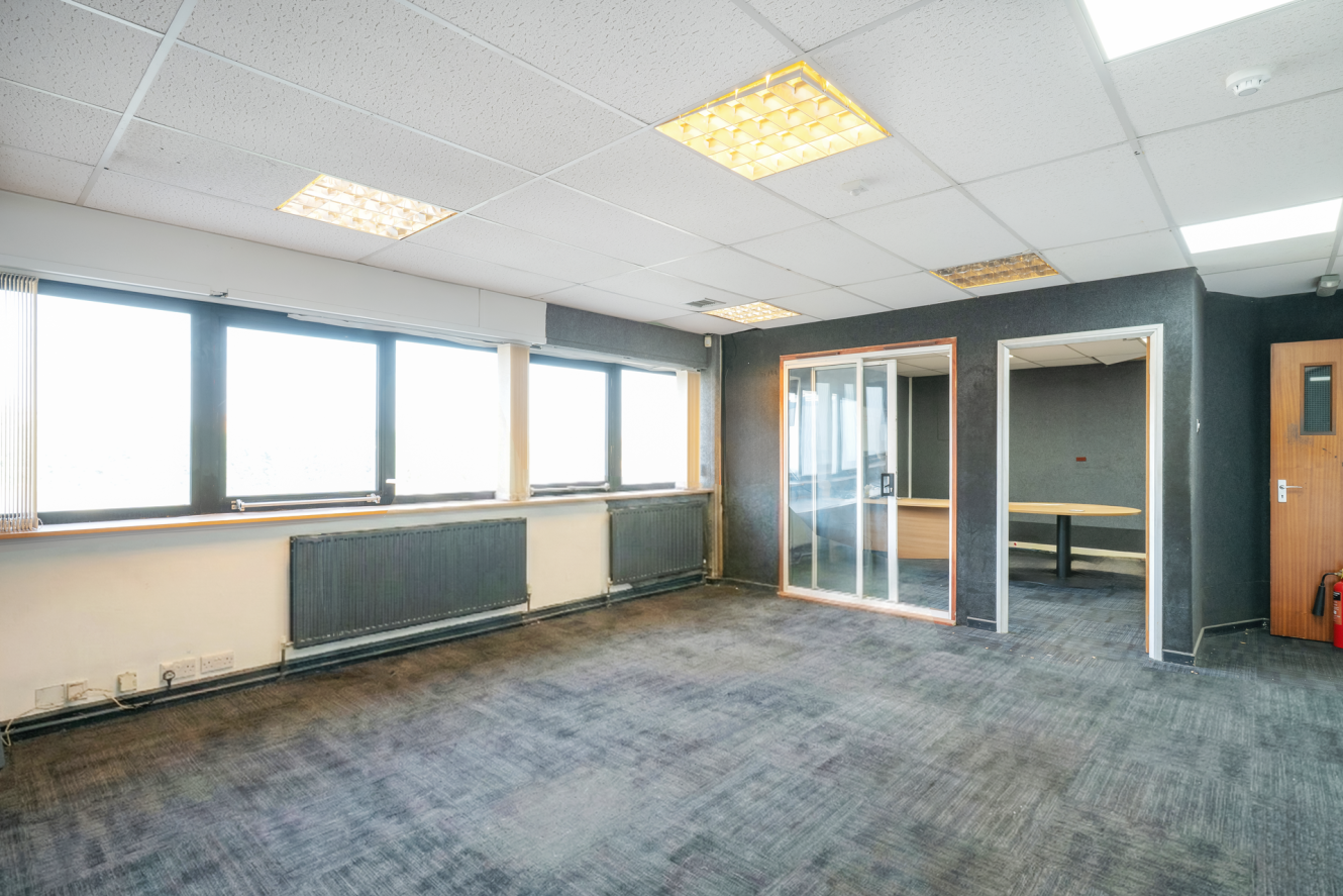 Empty office room with large windows, grey carpet, suspended ceiling with fluorescent lights, and a small glass-walled meeting room in the corner.