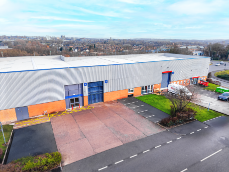 A large industrial warehouse with blue and red doors, a small car park, and surrounding greenery, located near a cityscape under a partly cloudy sky.