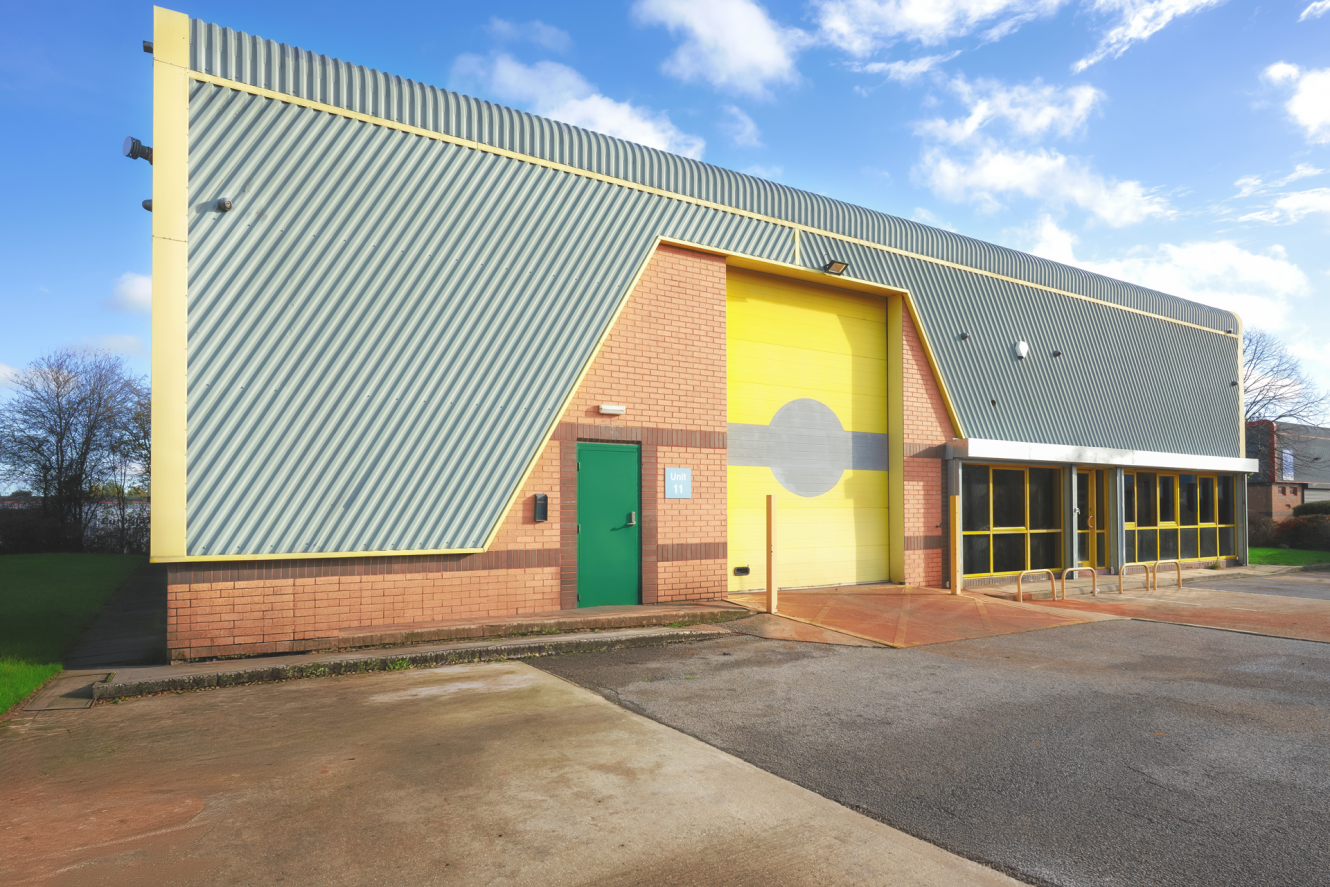 Industrial building with a green side door, large yellow garage door, brick lower walls, and corrugated metal cladding, with windows on the right side and a paved area in front.