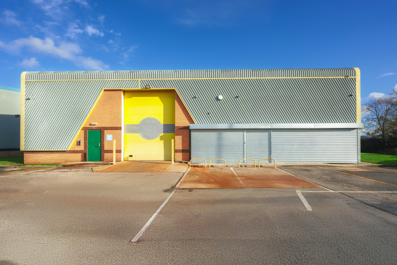 Industrial building with a yellow and green façade, metal garage doors, bicycle racks, and empty parking spaces under a clear blue sky.