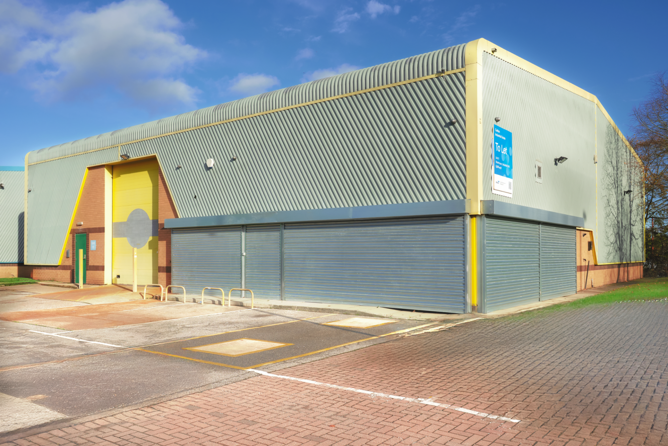 A light industrial warehouse with corrugated metal walls, a yellow door, closed roller shutters, and a “To Let” sign, situated next to a paved car park.