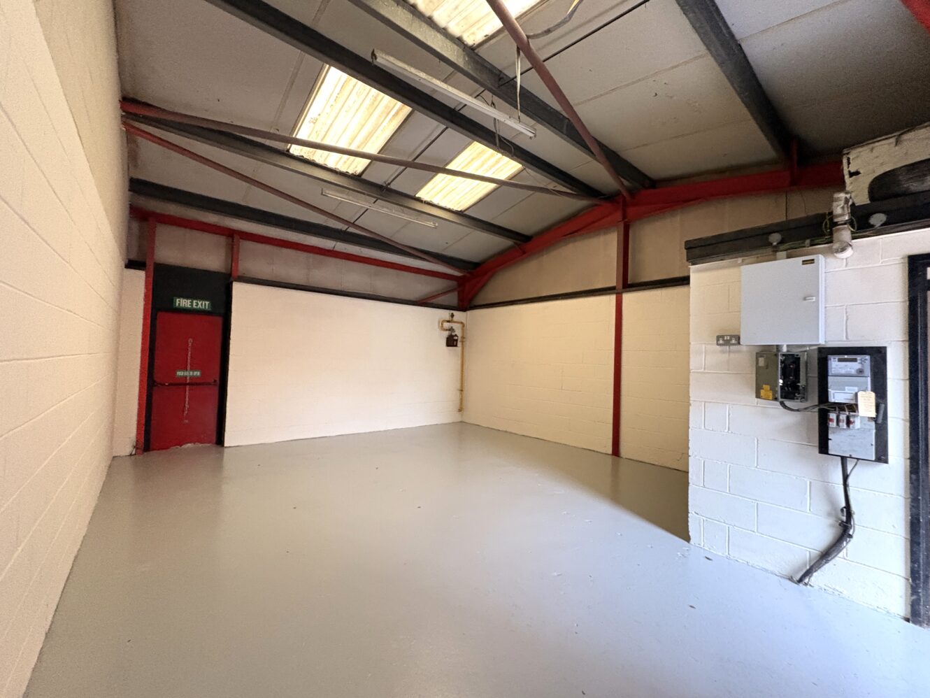 An empty industrial room with white walls, grey floor, red door labelled FIRE EXIT, exposed beams, and electrical panels on the right wall under fluorescent lights.