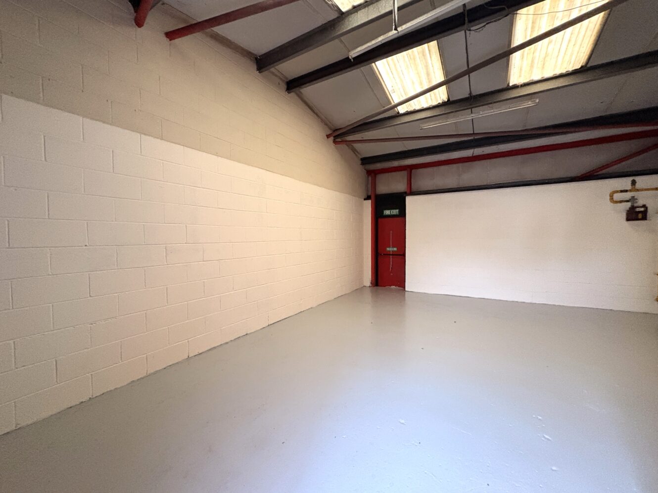 Empty industrial warehouse unit with white painted brick walls, grey concrete floor, exposed beams, and a red door at the back under fluorescent ceiling lights.