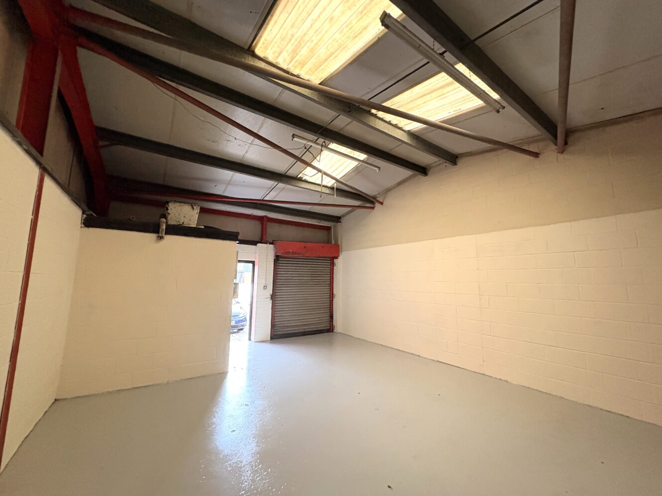 Empty industrial unit with white painted brick walls, a grey floor, exposed red beams, fluorescent lights, and a partially open roller shutter letting in daylight.