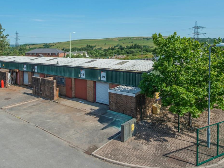 A row of industrial units with roller shutters, numbered 38 and 39, stands beside a tree on a sunny day with hills and power lines in the background.