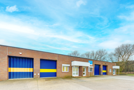 Single-storey industrial building with brick walls, blue metal doors featuring yellow stripes, small windows, and an overhang, set against a clear sky and bare trees.