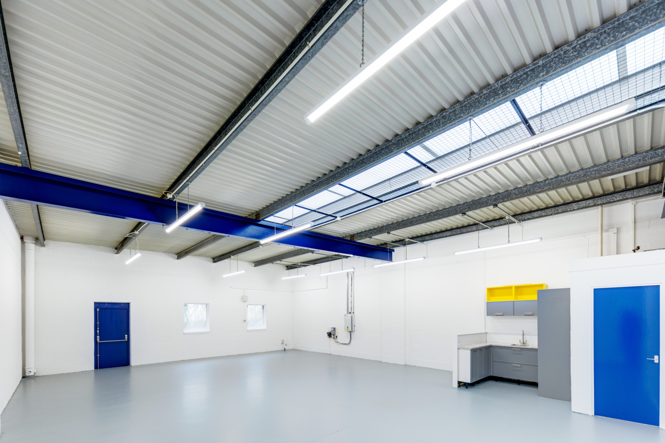 An empty industrial room with white walls, grey floor, fluorescent lights, exposed beams, two blue doors, two windows, and a kitchenette with yellow shelves.