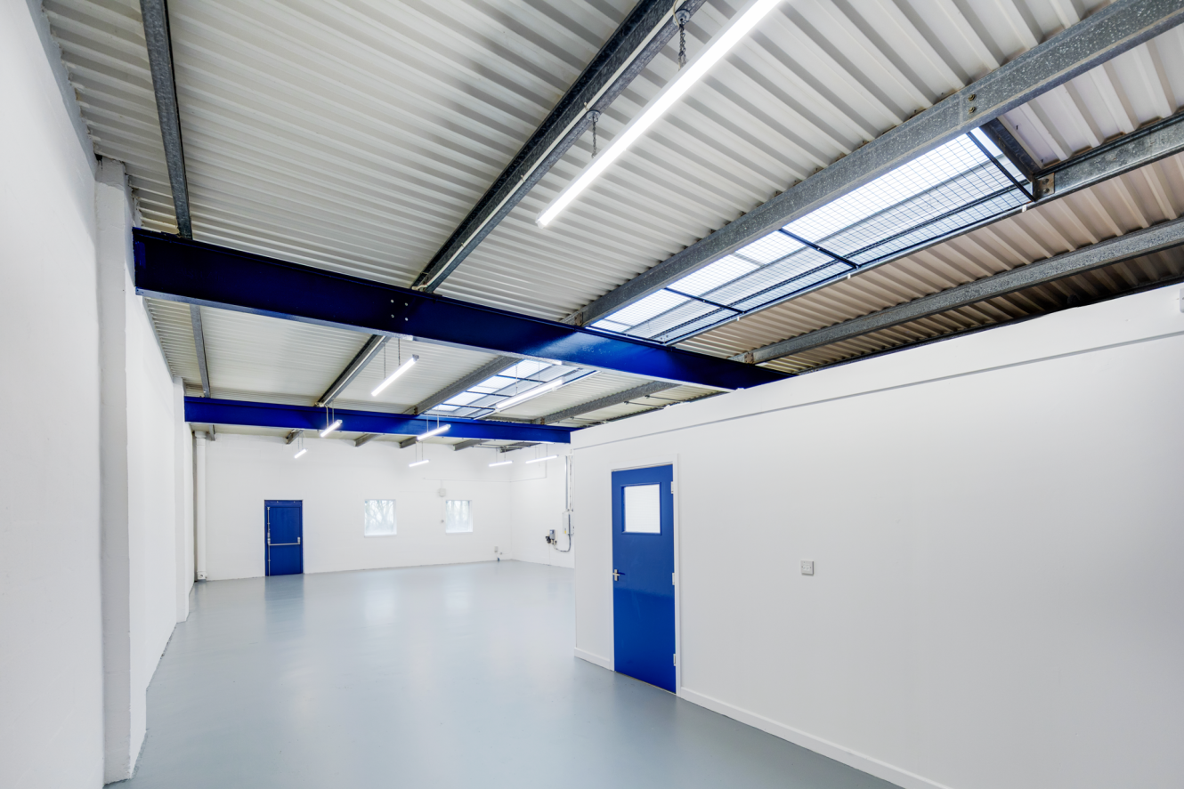 Interior of an empty industrial warehouse with white walls, grey floor, blue doors, exposed beams, and ceiling lights. Natural light enters through skylights.