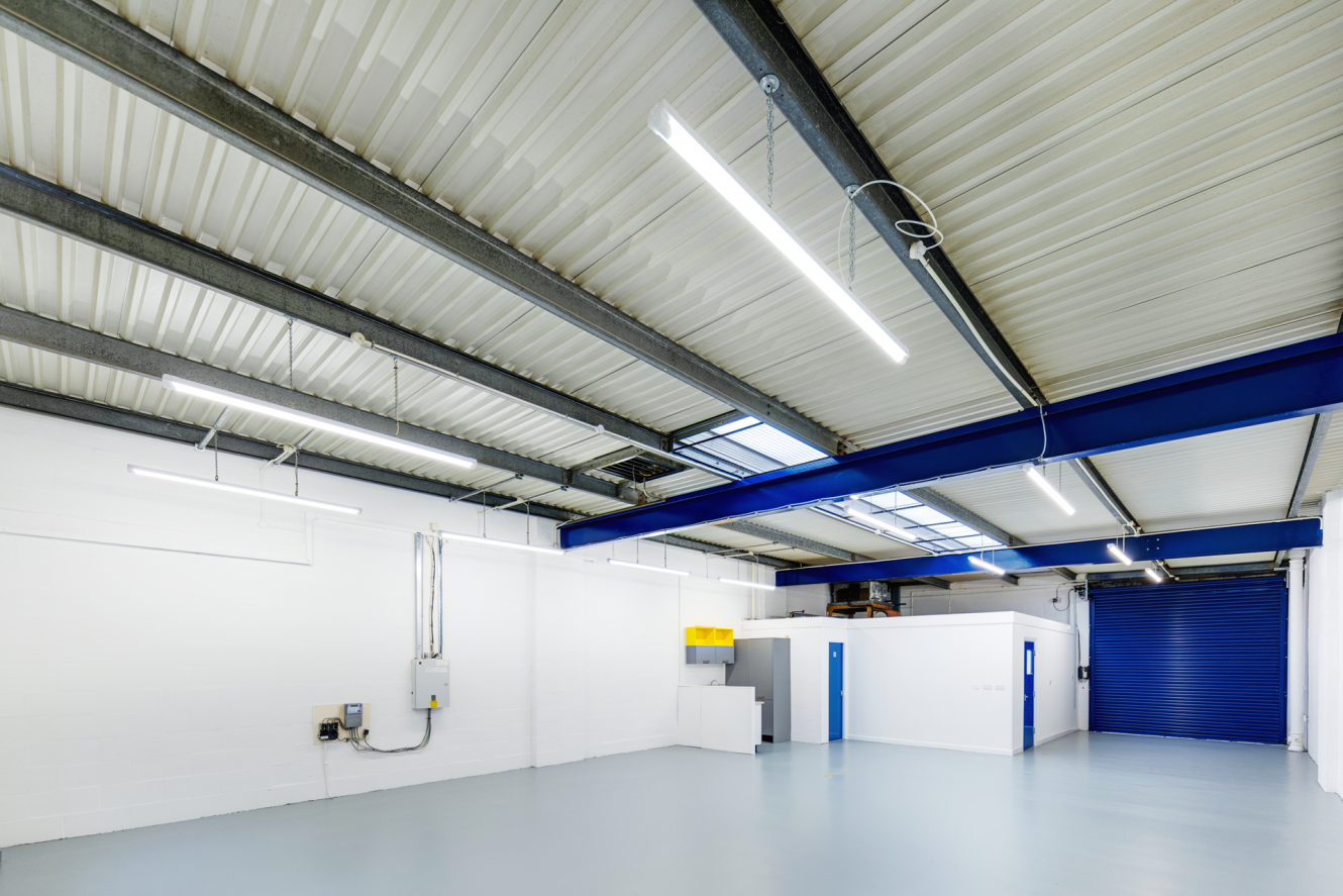 Interior of a clean, empty industrial warehouse with white walls, grey floor, exposed ceiling beams, fluorescent lighting, and a blue roller shutter door.