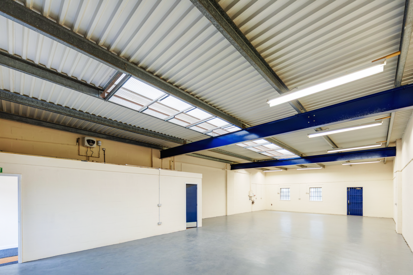 Empty industrial warehouse interior with white walls, exposed steel beams, fluorescent lighting, concrete floor, and two blue doors. Natural light enters through skylights and windows.