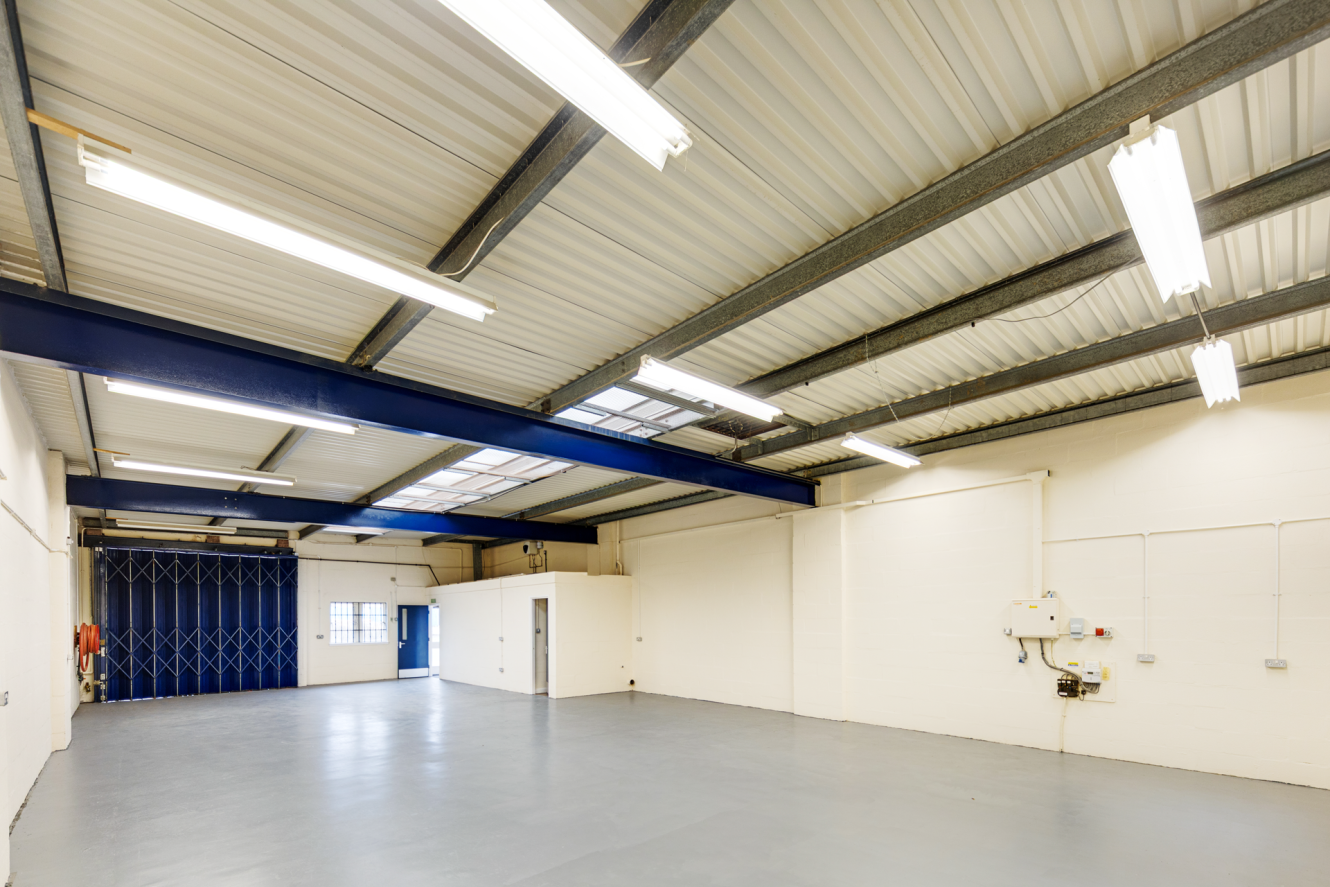 Empty industrial warehouse interior with white walls, grey concrete floor, overhead fluorescent lighting, and blue ceiling beams. A small office area and blue shutter doors are visible.
