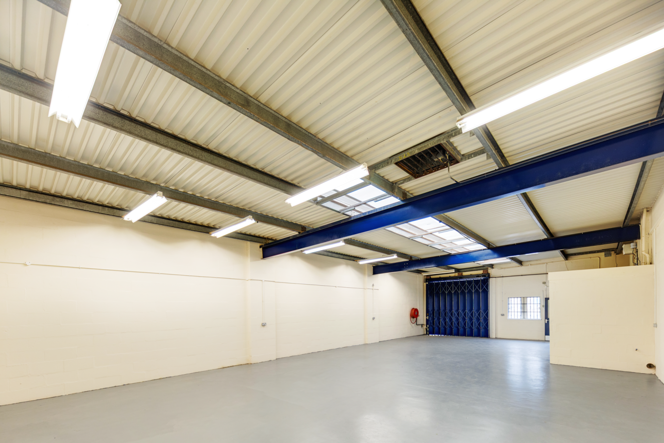 Empty industrial warehouse with white walls, concrete floor, fluorescent ceiling lights, and blue steel beams. A closed blue folding door and window are visible at the far end.