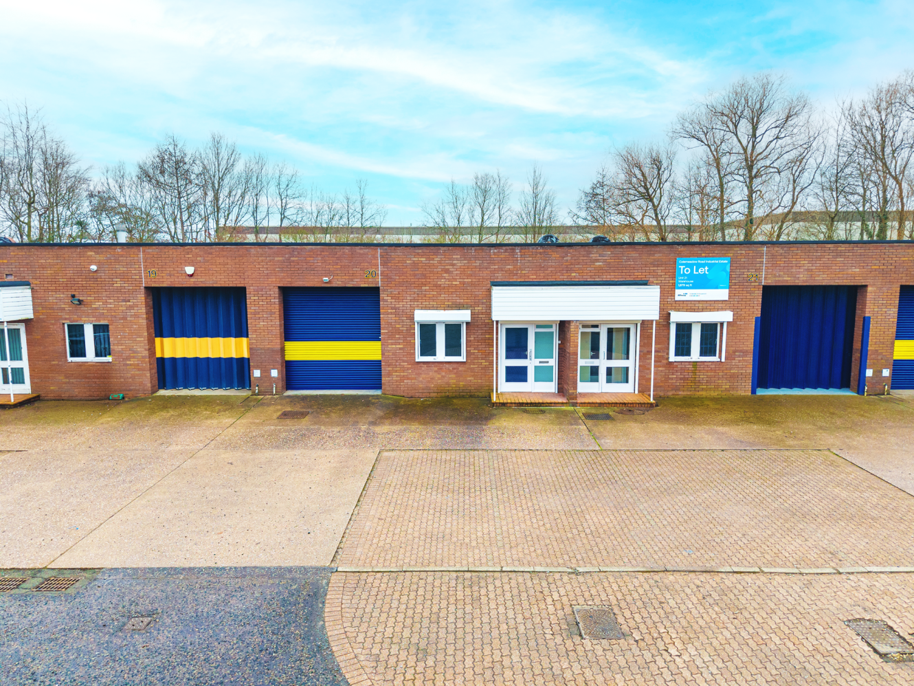 A row of brick industrial units with blue roller shutter doors, yellow stripes, and a To Let sign on one unit; empty car park in front.