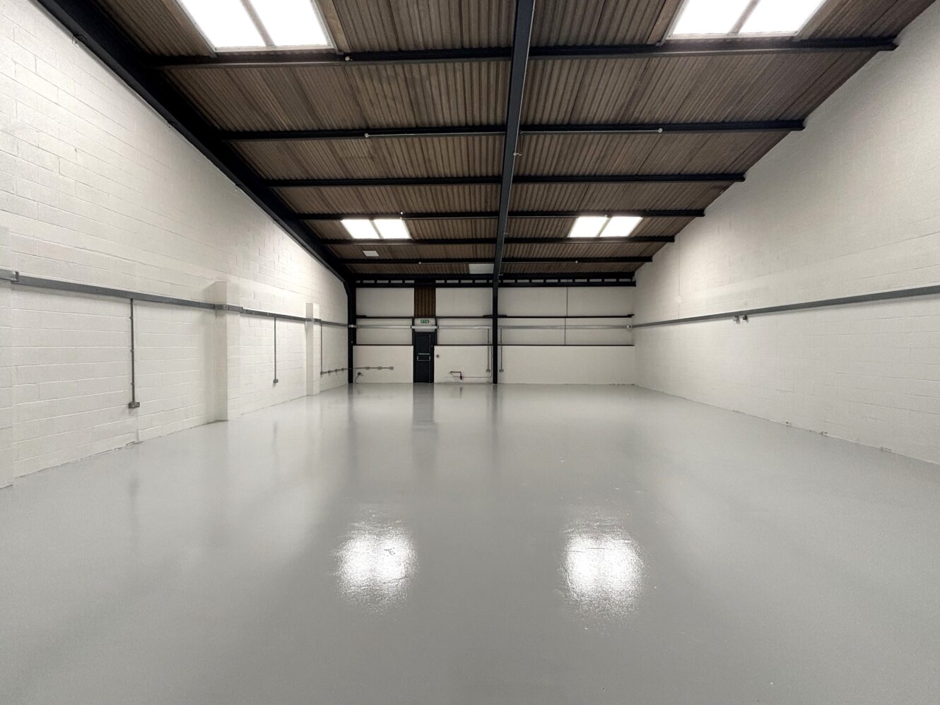 Empty industrial warehouse with white walls, high ceiling, and polished grey floor, lit by rectangular ceiling lights.