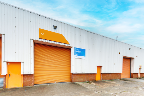 Industrial warehouse with white and orange exterior, large roller shutters, and a To Let sign displayed above one entrance.