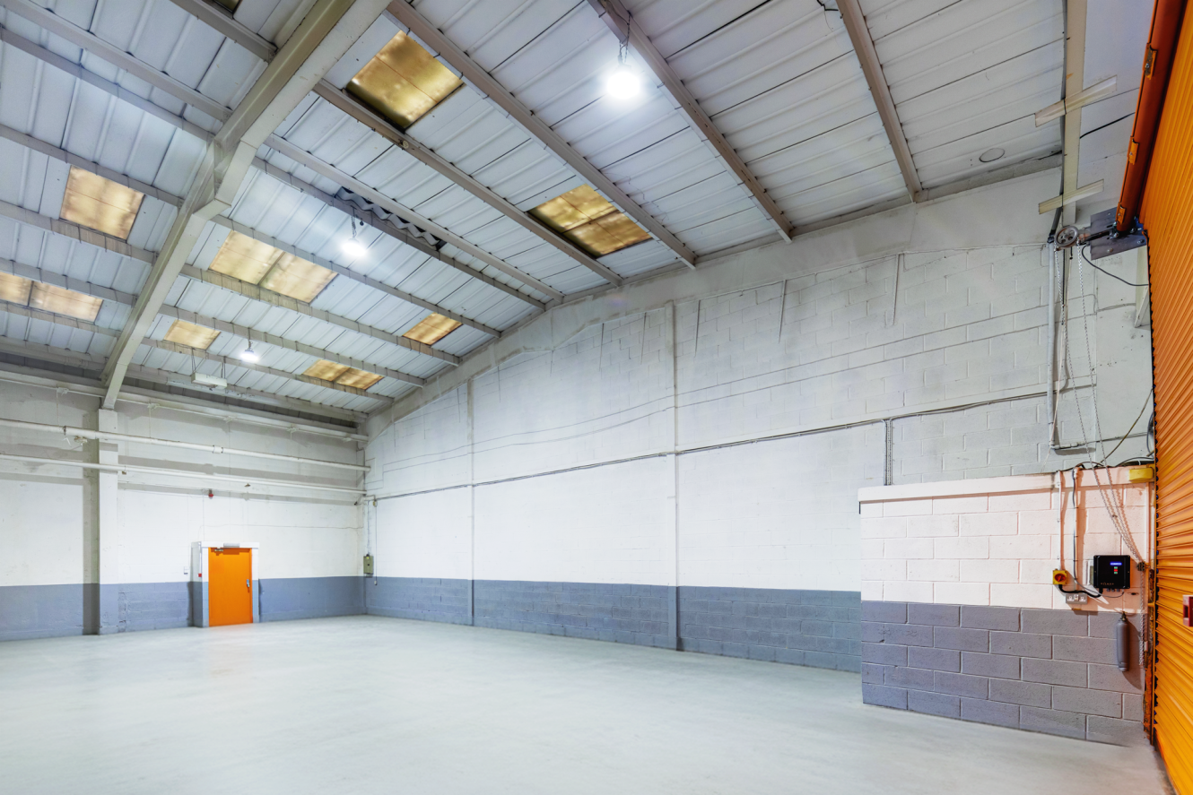 Interior of an empty industrial warehouse with high ceilings, concrete floor, white and grey walls, and two orange doors, lit by overhead lights.